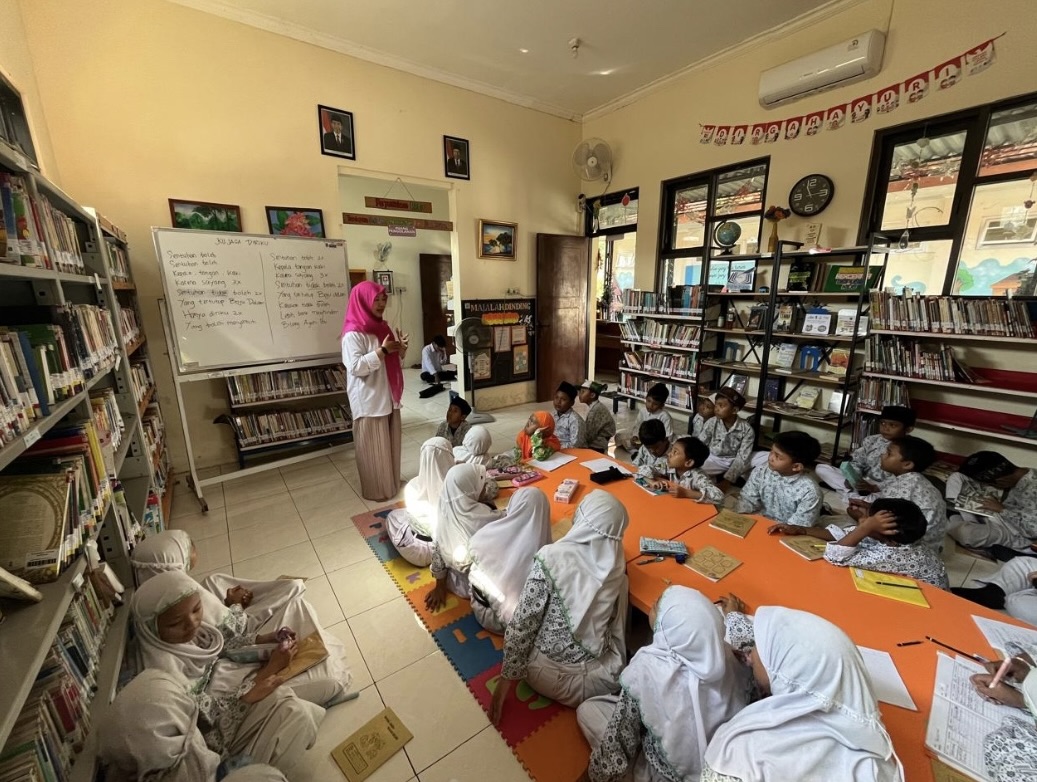 Children in white uniforms sitting on floor in library classroom with teacher in pink hijab